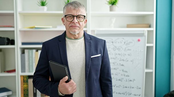 Hombre de pelo gris de mediana edad profesor de pie con expresión relajada sosteniendo touchpad en el aula universitaria - Foto, Imagen