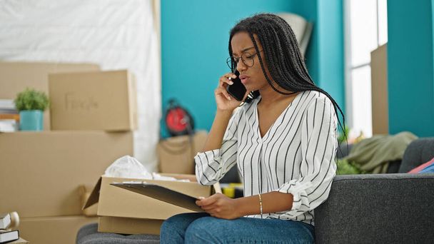 Mujer afroamericana hablando en un teléfono inteligente mirando molesto en un nuevo hogar - Foto, Imagen