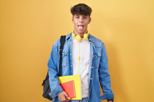 Hispanic teenager wearing student backpack and holding books sticking tongue out happy with funny expression. emotion concept.  - 写真・画像