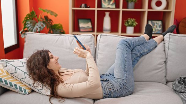 Mujer hispana hermosa joven usando teléfono inteligente tumbado en el sofá en casa - Foto, Imagen