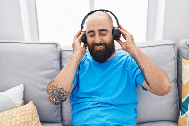 Young bald man listening to music sitting on sofa at home - Foto, Imagen