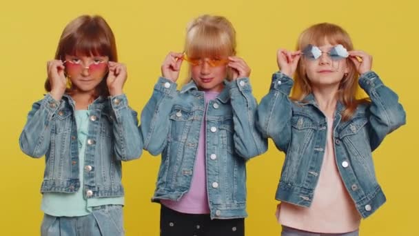 Teenage girls wearing sunglasses, charming smile. Little children sisters listening music, dancing disco fooling around having fun. Three siblings kids relaxing on party isolated on yellow background - Footage, Video