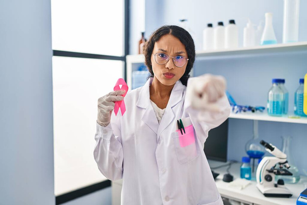 若いヒスパニックの医師女性は,カメラとあなたに指を指してピンクのリボンを保持する科学研究所で働いています,あなたに自信を持ってジェスチャーは深刻に見えます  - 写真・画像