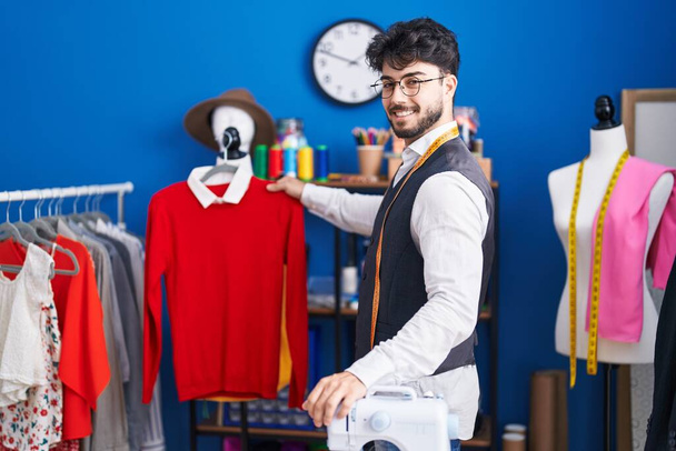 Young hispanic man tailor smiling confident holding clothes on rack at sewing studio - Valokuva, kuva