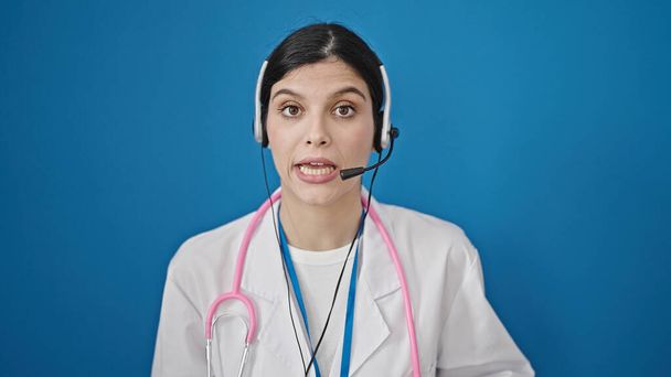 Junge schöne hispanische Ärztin lächelt zuversichtlich mit Online-ärztliche Beratung über isolierten blauen Hintergrund - Foto, Bild