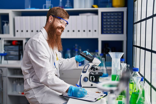 Νεαρή κοκκινομάλλα επιστήμονας χρησιμοποιώντας μικροσκόπιο γραφής έκθεση εργασίας στο εργαστήριο - Φωτογραφία, εικόνα