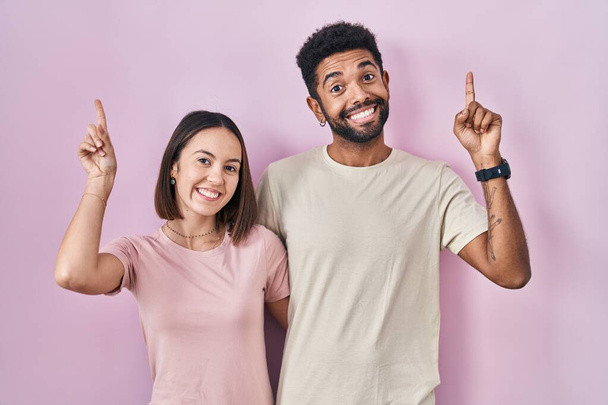 Jeune couple hispanique ensemble sur fond rose sourire étonné et surpris et pointant vers le haut avec les doigts et les bras levés.  - Photo, image