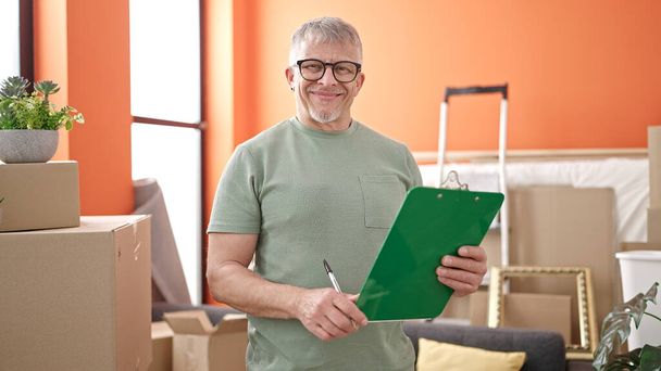 Hombre de pelo gris de mediana edad sonriendo confiado sujetando el portapapeles en un nuevo hogar - Foto, Imagen