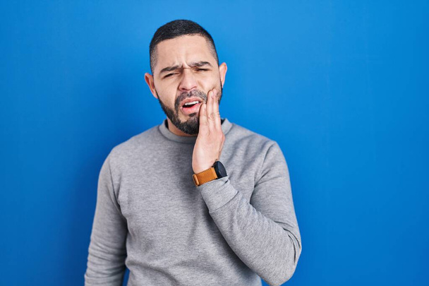 Uomo ispanico in piedi sopra sfondo blu toccare bocca con mano con espressione dolorosa a causa di mal di denti o malattia dentale sui denti. dentista  - Foto, immagini