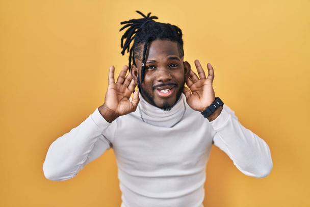 African man with dreadlocks wearing turtleneck sweater over yellow background trying to hear both hands on ear gesture, curious for gossip. hearing problem, deaf  - Фото, изображение