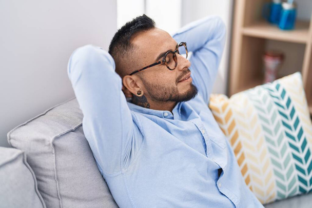Jeune homme hispanique détendu avec les mains sur la tête assis sur le canapé à la maison - Photo, image