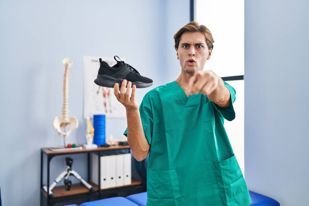Young man working at physiotherapy clinic holding shoe pointing with finger to the camera and to you, confident gesture looking serious  - Photo, Image