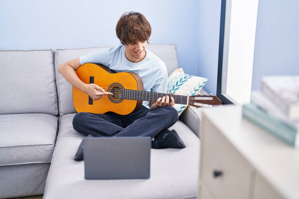 Jonge blonde man met online klassieke gitaar klasse zitten op de bank thuis - Foto, afbeelding