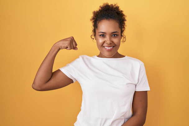 Young hispanic woman with curly hair standing over yellow background strong person showing arm muscle, confident and proud of power  - Photo, Image