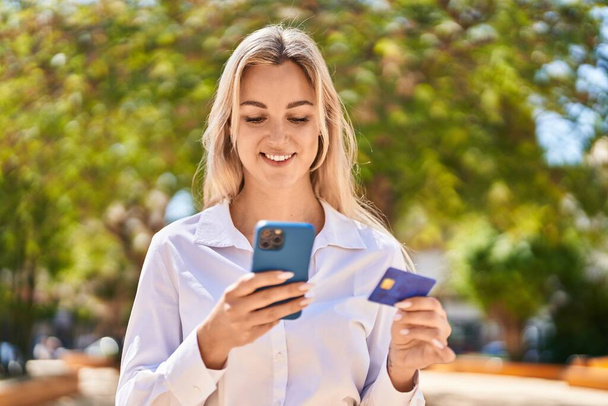 Jonge blonde vrouw met smartphone en creditcard in het park - Foto, afbeelding