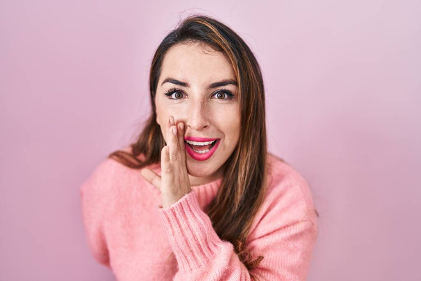 Fiatal spanyol nő áll a rózsaszín háttér kéz a szájban mondja a titkos pletyka, suttogó rosszindulatú beszélgetés  - Fotó, kép