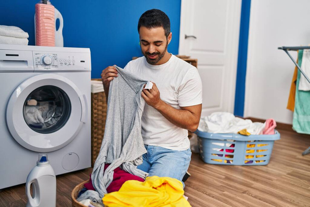 Giovane uomo ispanico lavare i vestiti in cerca di etichetta t shirt in lavanderia - Foto, immagini