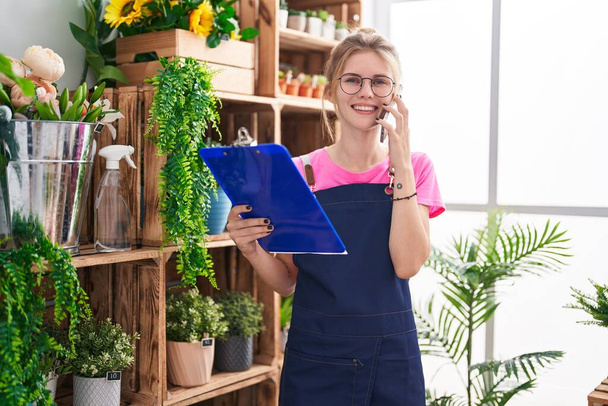Young blonde woman florist talking on smartphone reading clipboard at flower shop - Photo, Image