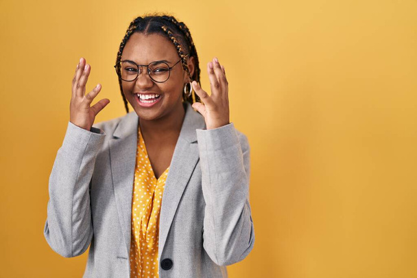 Femme afro-américaine avec des tresses debout sur fond jaune célébrant fou et étonné pour le succès avec les bras levés et les yeux ouverts hurlant excité. concept gagnant  - Photo, image
