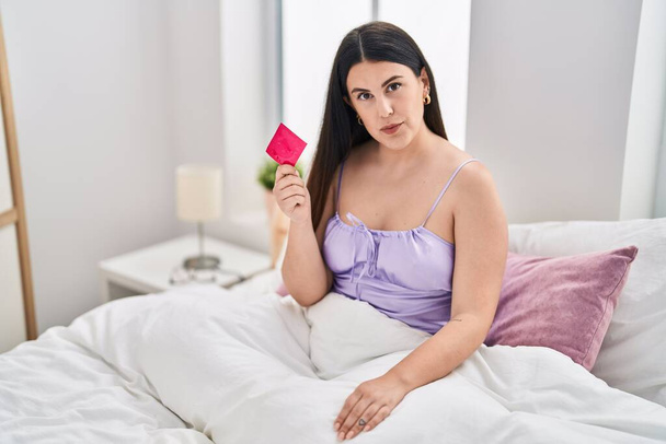Giovane donna bruna che indossa lingerie e tiene il preservativo sul letto pensando atteggiamento ed espressione sobria guardando fiducioso di sé  - Foto, immagini
