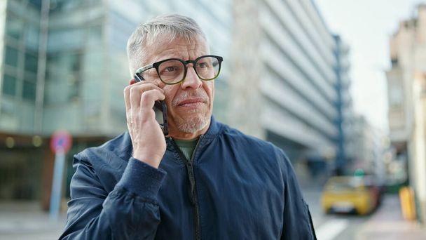 Moyen-âge homme aux cheveux gris parler sur smartphone avec expression inquiète à la rue - Photo, image