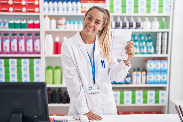 Молодая красивая латиноамериканка-фармацевт улыбается уверенно держа рецептурную бумагу в аптеке - Фото, изображение