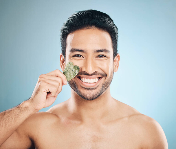 Face, man and beauty, gua sha and cosmetic tools, smile in portrait and natural treatment on blue background. Skincare, healthy glow and jade stone, dermatology and wellness with male model in studio. - Photo, Image