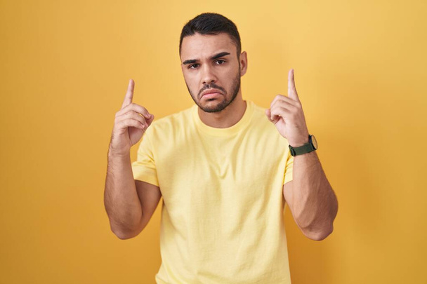 Junger hispanischer Mann, der vor gelbem Hintergrund steht und traurig und aufgebracht nach oben zeigt, mit den Fingern Richtung zeigt, unglücklich und deprimiert.  - Foto, Bild