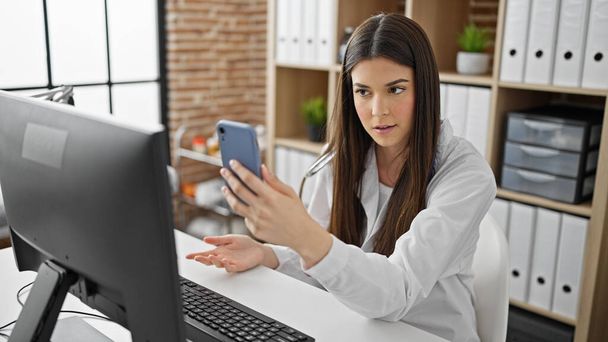 Genç, güzel İspanyol kadın doktor klinikte akıllı telefonuyla video görüşmesi yapıyor. - Fotoğraf, Görsel