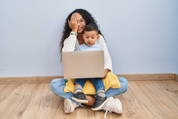 Νέοι Ισπανοί μητέρα και το παιδί χρησιμοποιώντας φορητό υπολογιστή κάθεται στο πάτωμα καλύπτει το ένα μάτι με το χέρι, αυτοπεποίθηση χαμόγελο στο πρόσωπο και έκπληξη συναίσθημα.  - Φωτογραφία, εικόνα