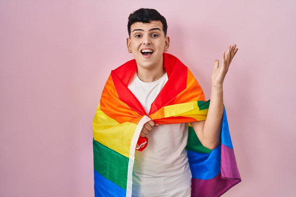 Persona no binaria sosteniendo la bandera del arco iris lgbtq celebrando la victoria con sonrisa feliz y expresión ganadora con las manos levantadas  - Foto, imagen