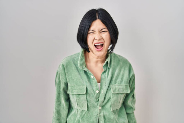 Young asian woman standing over white background angry and mad screaming frustrated and furious, shouting with anger. rage and aggressive concept.  - Fotó, kép