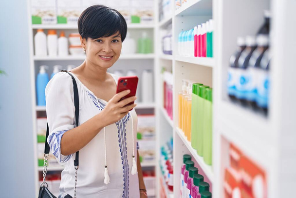 Середній вік жінка - клієнтка посміхається впевнено, використовуючи смартфон в аптеці. - Фото, зображення