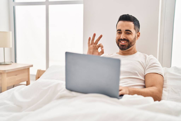 Νεαρός Ισπανόφωνος με γενειάδα χρησιμοποιώντας φορητό υπολογιστή στο κρεβάτι κάνει ok υπογράψει με τα δάχτυλα, χαμογελώντας φιλικό gesturing εξαιρετικό σύμβολο  - Φωτογραφία, εικόνα
