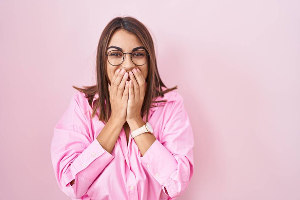 Νεαρή Ισπανίδα γυναίκα με γυαλιά που στέκεται πάνω από ροζ φόντο γελώντας και ντροπιασμένη χαχανίζοντας καλύπτοντας το στόμα με χέρια, κουτσομπολιά και σκανδαλώδη έννοια  - Φωτογραφία, εικόνα