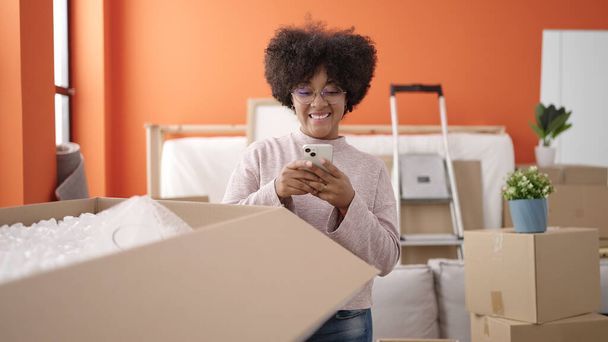 Mujer afroamericana joven desempaquetando la caja de cartón usando el teléfono inteligente en el nuevo hogar - Foto, imagen