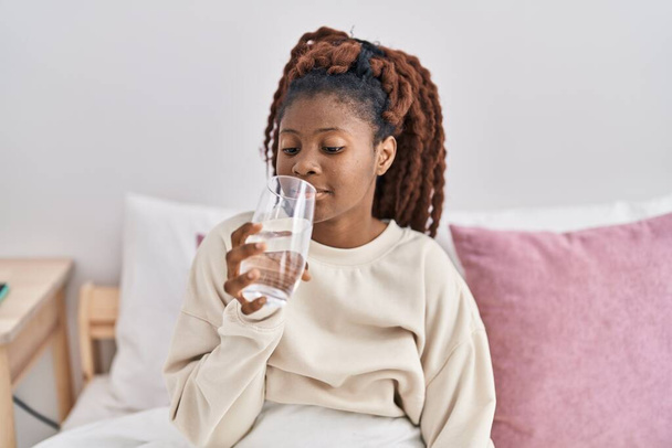 African american woman drinking glass of water sitting on bed at bedroom - Foto, Imagem
