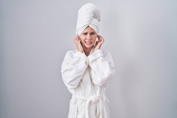 Blonde caucasian woman wearing bathrobe covering ears with fingers with annoyed expression for the noise of loud music. deaf concept.  - Foto, Imagen