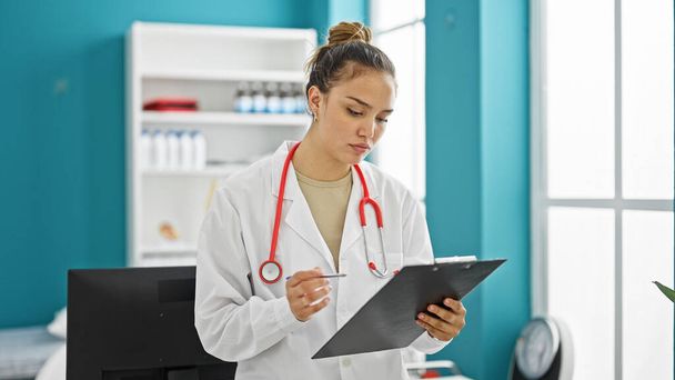 Joven hermosa mujer hispana médico leyendo documento en portapapeles con cara seria en la clínica - Foto, Imagen