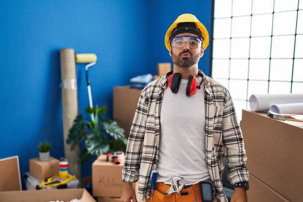 Ein junger hispanischer Mann mit Bart, der zu Hause Renovierungsarbeiten verrichtet und in die Kamera schaut, während er einen Kuss in die Luft pustet, ist schön und sexy. Liebesausdruck.  - Foto, Bild
