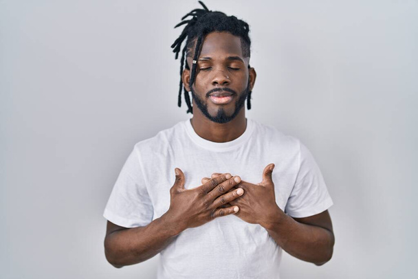 Африканский мужчина с дредами в повседневной футболке на белом фоне улыбается руками на груди с закрытыми глазами и благодарным жестом на лице. концепция здоровья.  - Фото, изображение