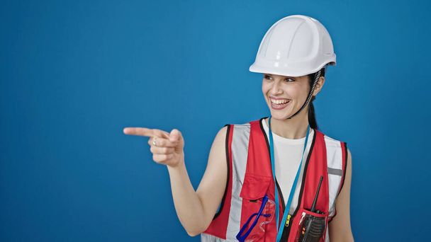 Молода красива іспаномовна жінка-будівельник посміхається впевнено вказуючи на бік над ізольованим синім тлом
 - Фото, зображення