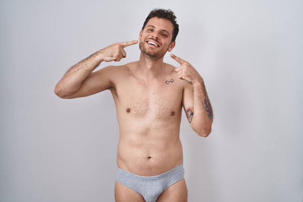 Jeune homme hispanique debout torse nu portant des sous-vêtements souriant gai montrant et pointant avec les doigts les dents et la bouche. concept de santé dentaire.  - Photo, image