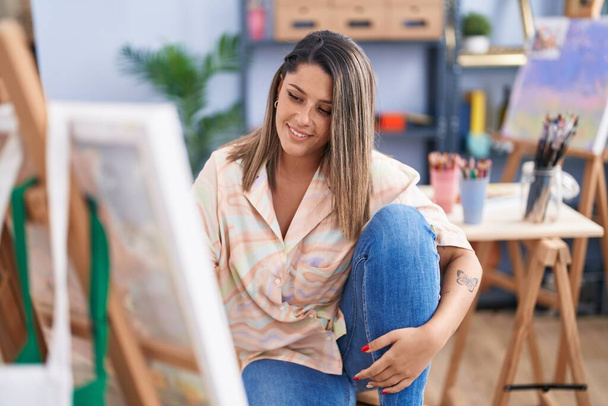 Młoda Latynoska artystka uśmiecha się pewnie siedząc na podłodze w pracowni sztuki - Zdjęcie, obraz