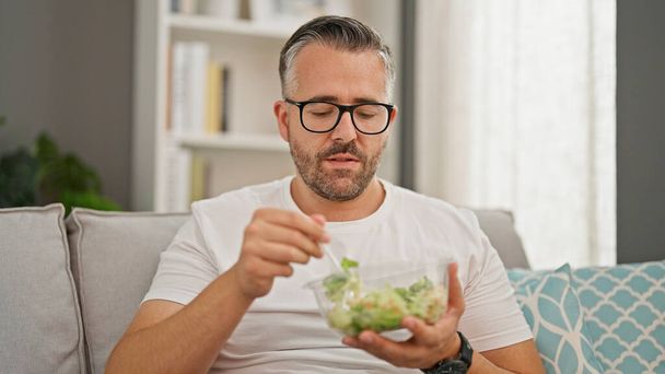 Homme aux cheveux gris mangeant de la salade assis sur le canapé à la maison - Photo, image