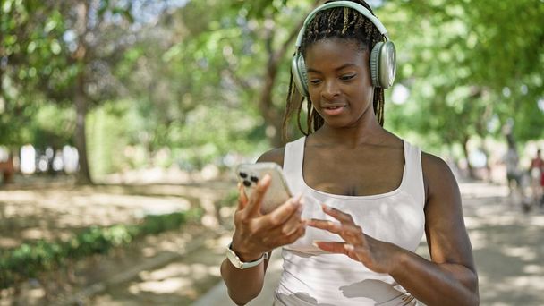 公園でスマートフォンやヘッドフォンを使うアフリカ系アメリカ人女性 - 写真・画像