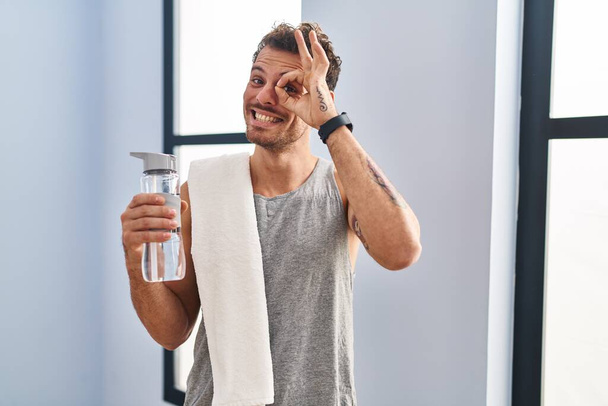 Jeune homme hispanique portant des vêtements de sport boire de l'eau faisant un geste correct avec la main souriante, les yeux regardant à travers les doigts avec un visage heureux.  - Photo, image