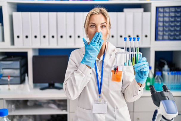 Jonge blanke vrouw die in het laboratorium werkt en reageerbuisjes vasthoudt die mond met de hand bedekken, geschokt en bang voor fouten. verraste uitdrukking  - Foto, afbeelding