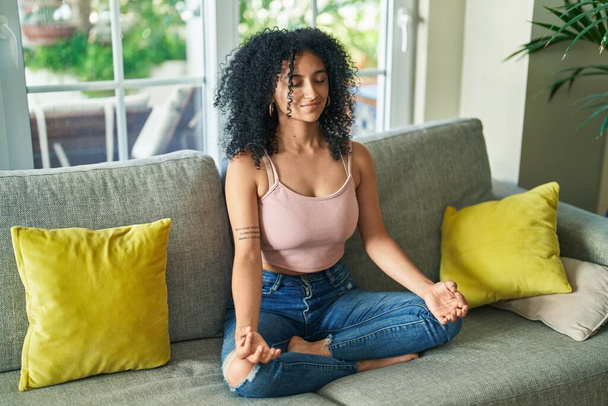 Jeune femme hispanique faisant de l'exercice de yoga assis sur le canapé à la maison - Photo, image