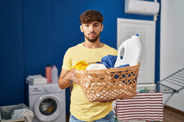 Arab man with beard holding laundry basket and detergent bottle skeptic and nervous, frowning upset because of problem. negative person.  - Фото, изображение